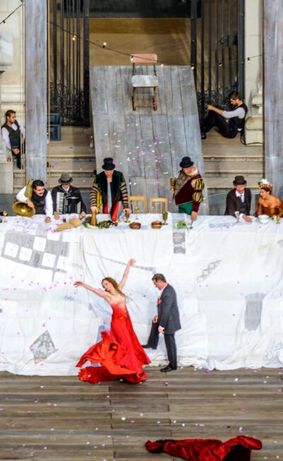 A dramatic outdoor performance at the Salzburg Festival features costumed actors, musicians, and a dancer in red.