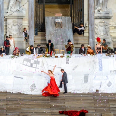A dramatic outdoor performance at the Salzburg Festival features costumed actors, musicians, and a dancer in red.