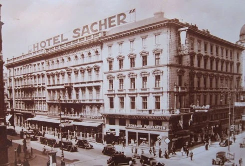A historic photograph of Hotel Sacher captures bustling street life and vintage cars in front of the iconic building.
