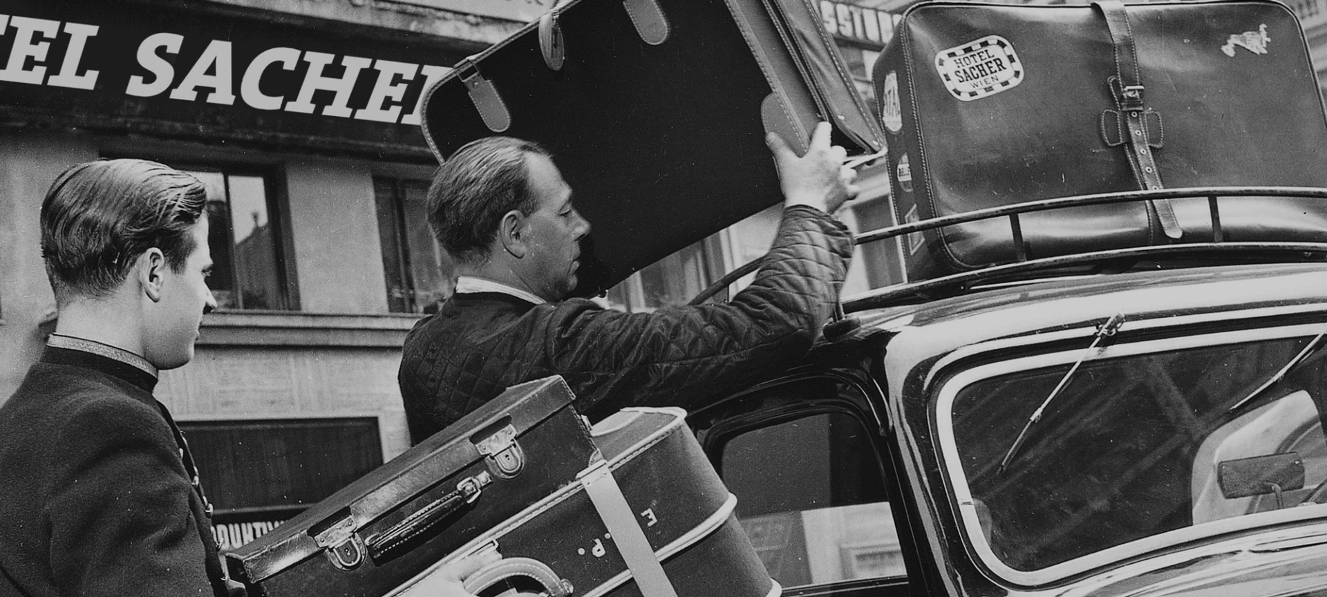 Black and white image of two Hotel Sacher valets putting suitcases onto the top of a car in front of the iconic hotel.