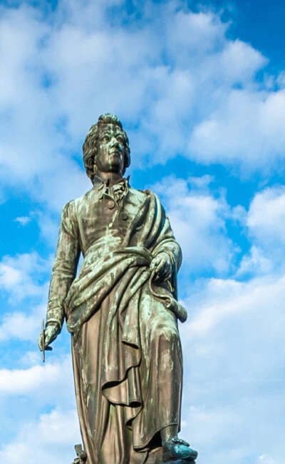 The image shows the Mozart monument in Salzburg, Austria, set against a vivid blue sky with scattered clouds.
