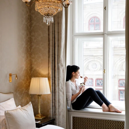 A woman enjoys a dessert while sitting on a windowsill, basking in the cozy elegance of her room with a view of a grand historic building.