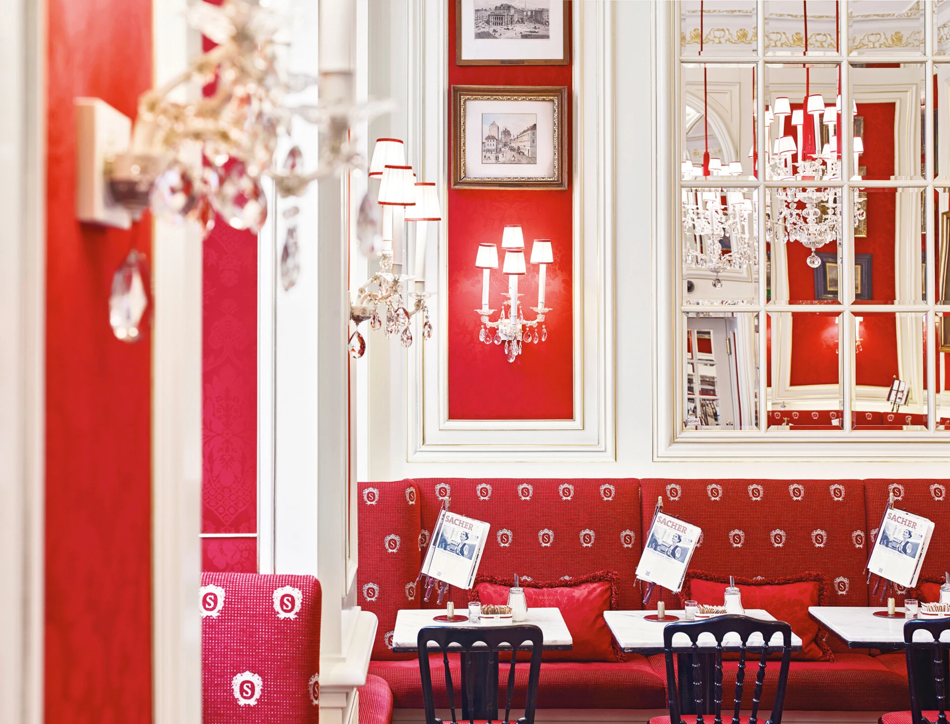 A charming view of Café Sacher, featuring elegant red-and-white decor, crystal chandeliers, classic framed art, and plush seating adorned with the iconic 