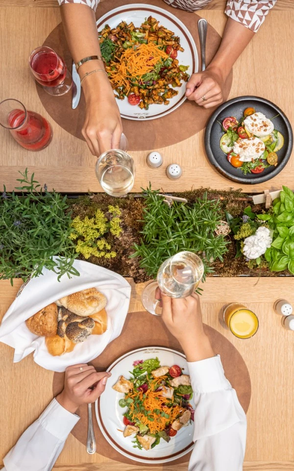A vibrant overhead view of two people clinking glasses over a healthy meal featuring colorful salads, fresh bread, and drinks on a beautifully set table with a green centerpiece.