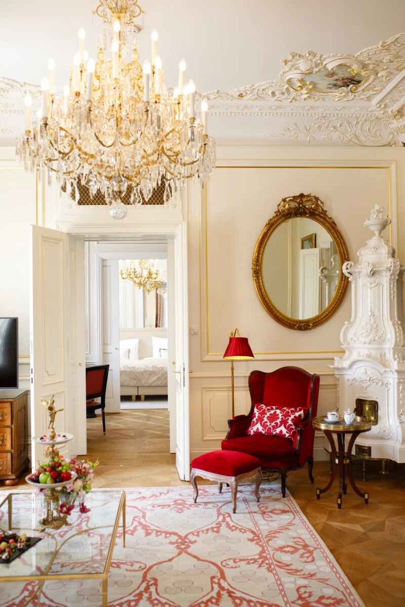 A luxurious Viennese suite features elegant chandeliers, ornate decor, and a plush red armchair beside a white ceramic stove.