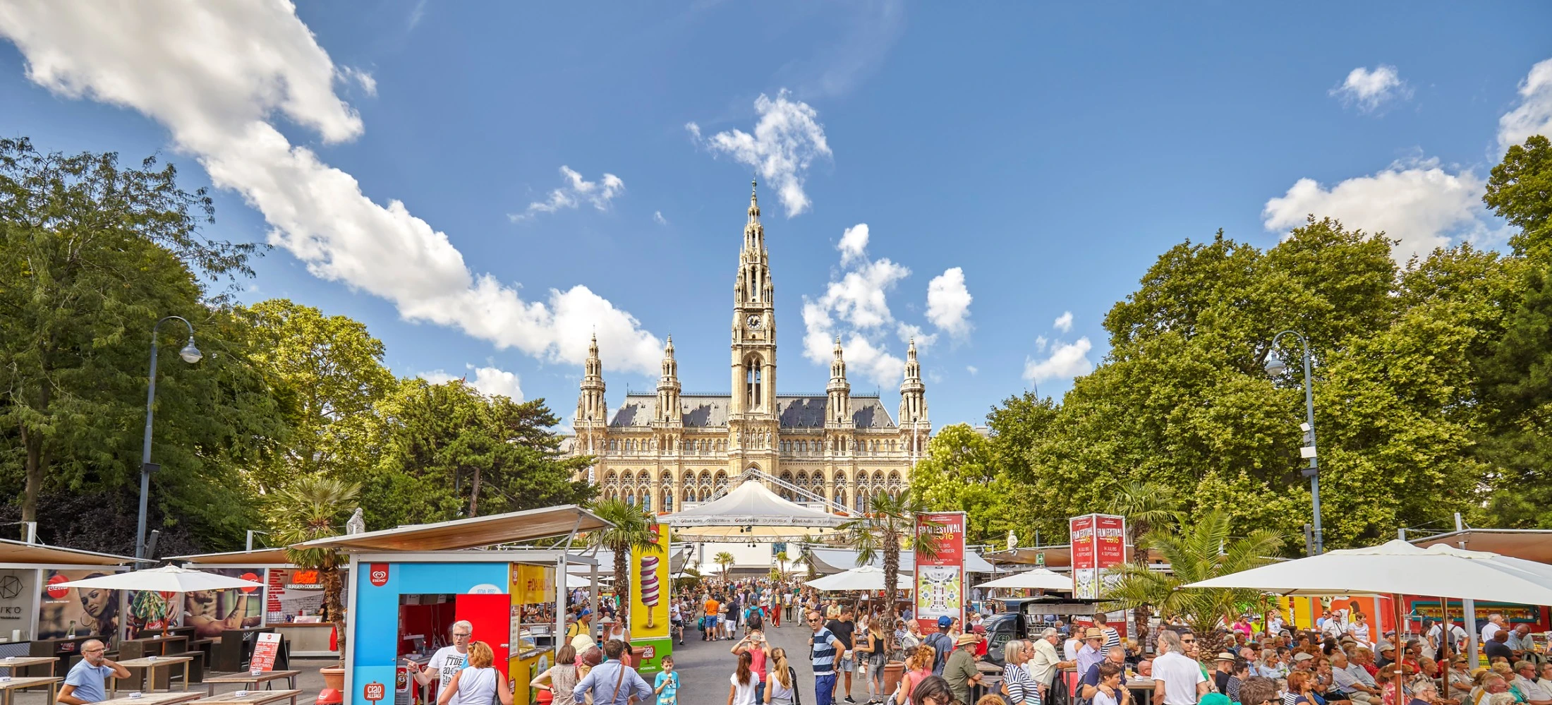Crowds enjoy a summer festival in front of Vienna’s grand Rathaus under a clear blue sky.