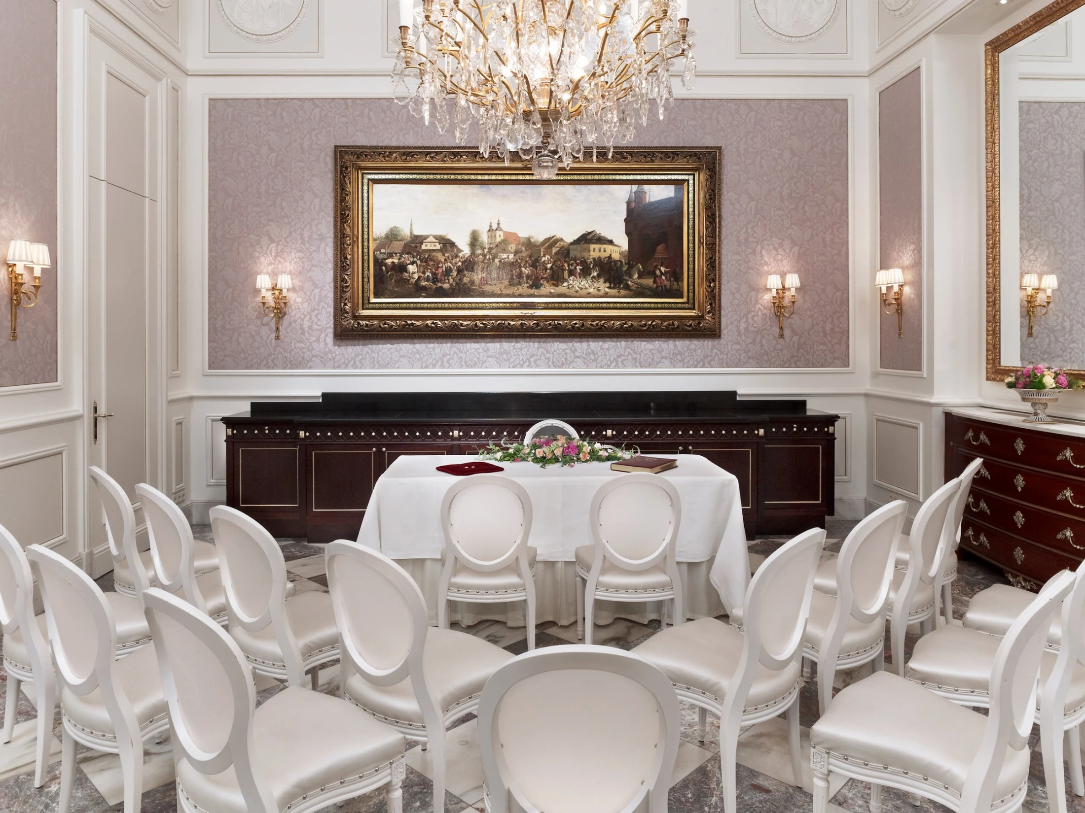 An elegant wedding ceremony setup in a classical room with white chairs, a chandelier, and floral decorations.