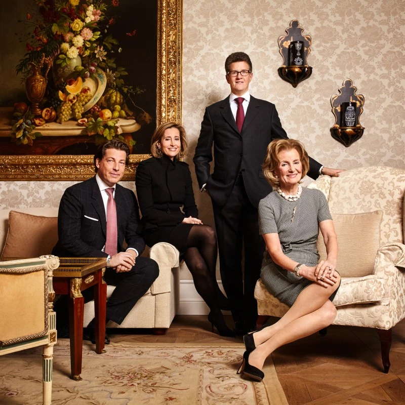 Elegant portrait of four individuals – two men in suits and two women in dresses – posed in a richly decorated, classic interior with an ornate still-life painting and antique furniture.