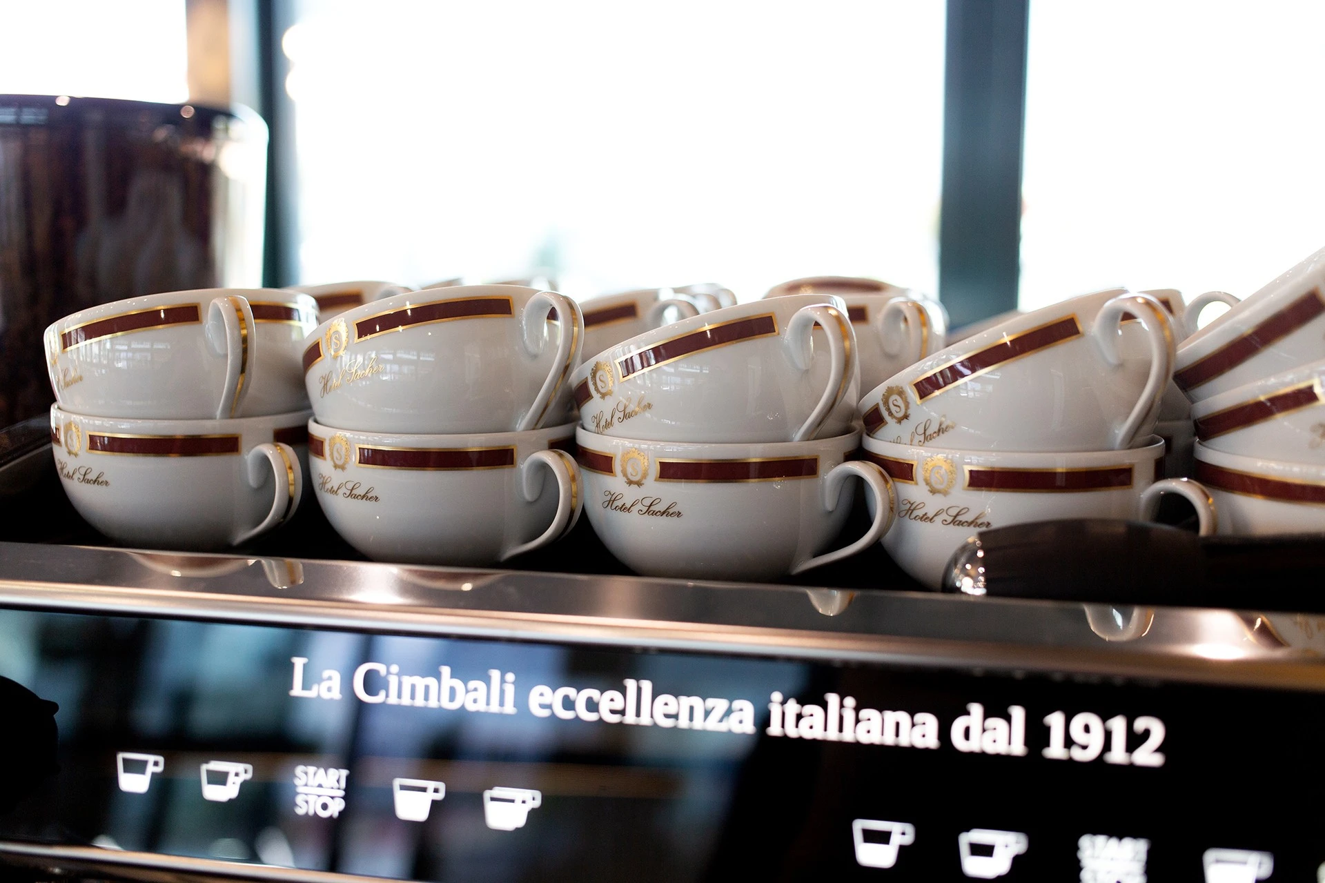 Stacks of elegant Hotel Sacher cups rest atop a La Cimbali espresso machine, promising an authentic and refined coffee experience.