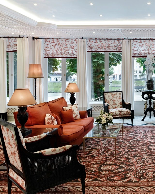 A classic lounge with views of the Salzach River, featuring rich red upholstery, elegant floral patterns, and black lacquered accents.