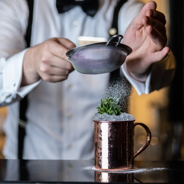 A bartender in formal attire is dusting powdered sugar over a cocktail served in a copper mug filled with crushed ice and mint.