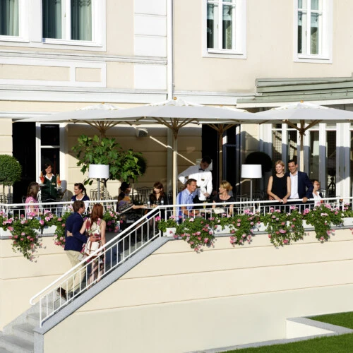 Guests are enjoying drinks and conversation on a flower-adorned terrace at Hotel Sacher Salzburg under large white umbrellas.
