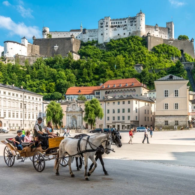 This image captures a classic horse-drawn carriage ride in Salzburg’s historic center, with Hohensalzburg Fortress towering above the city and lush greenery on the hillside.