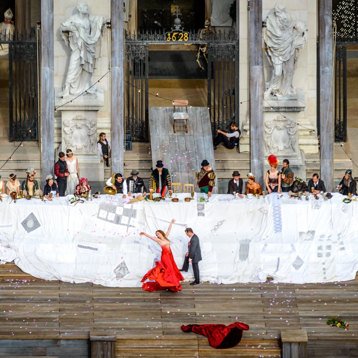 This image captures a dramatic outdoor performance at the Salzburger Festspiele, with actors in period costumes gathered at a long banquet table and a performer in red dancing on the stage below.