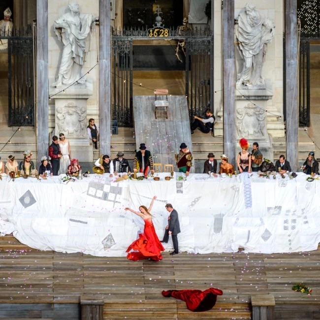 This image captures a dramatic outdoor performance at the Salzburger Festspiele, with actors in period costumes gathered at a long banquet table and a performer in red dancing on the stage below.
