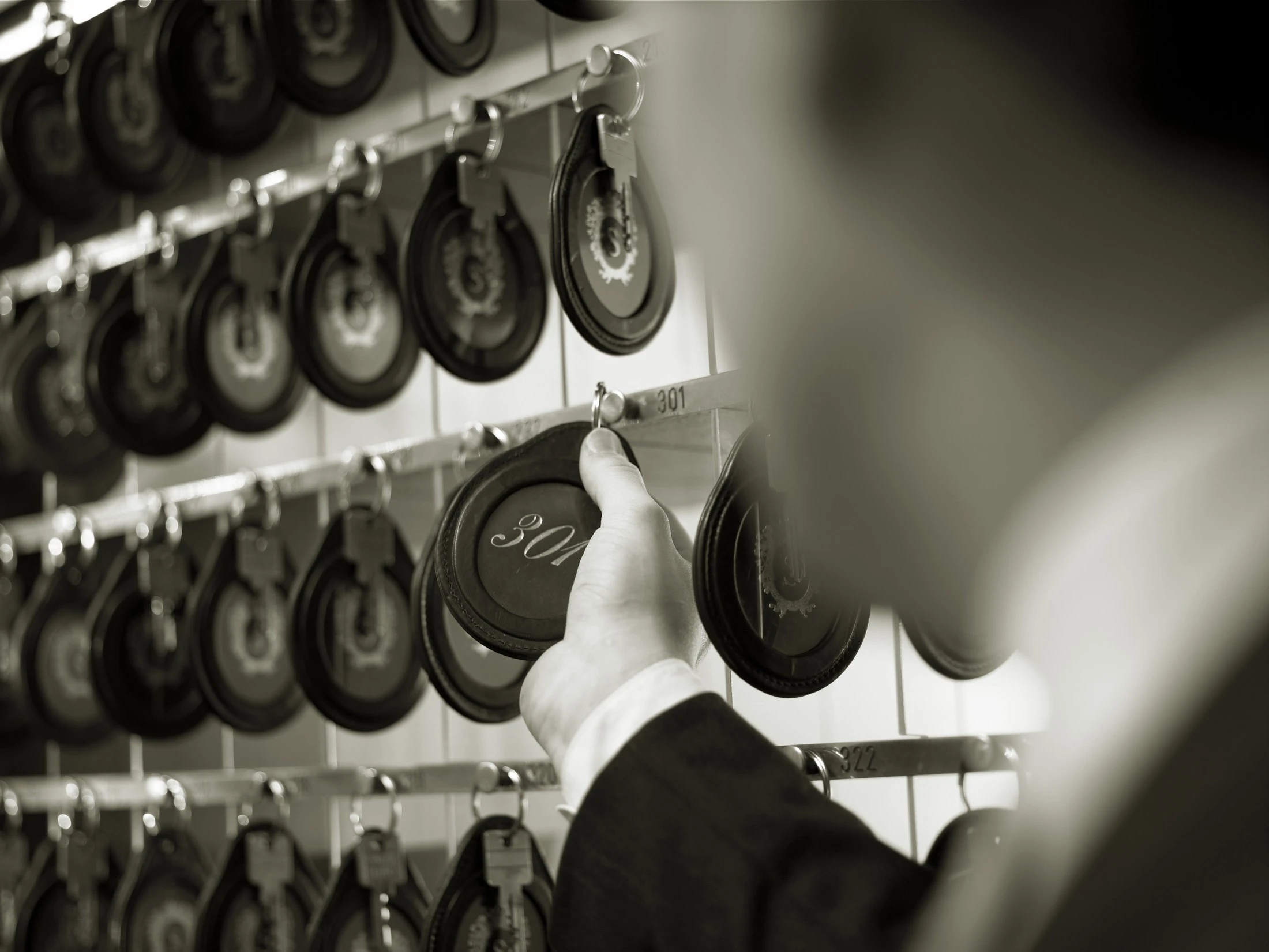 This black-and-white image shows a hotel staff member selecting a vintage room key labeled “301” from a wall of similar keys, evoking classic hospitality charm.