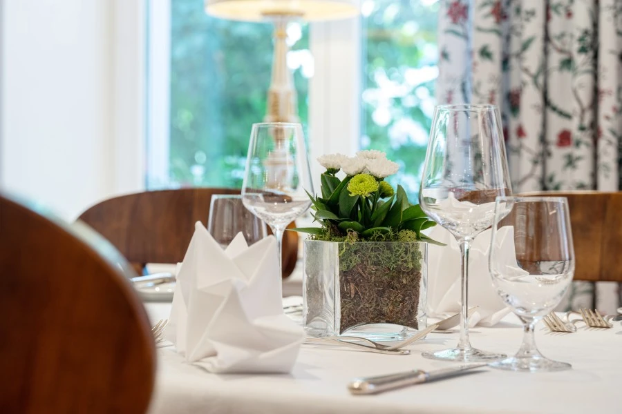 This image shows a close-up of a neatly set table with white napkins, wine glasses, and a small floral centerpiece in a clear glass vase filled with moss.