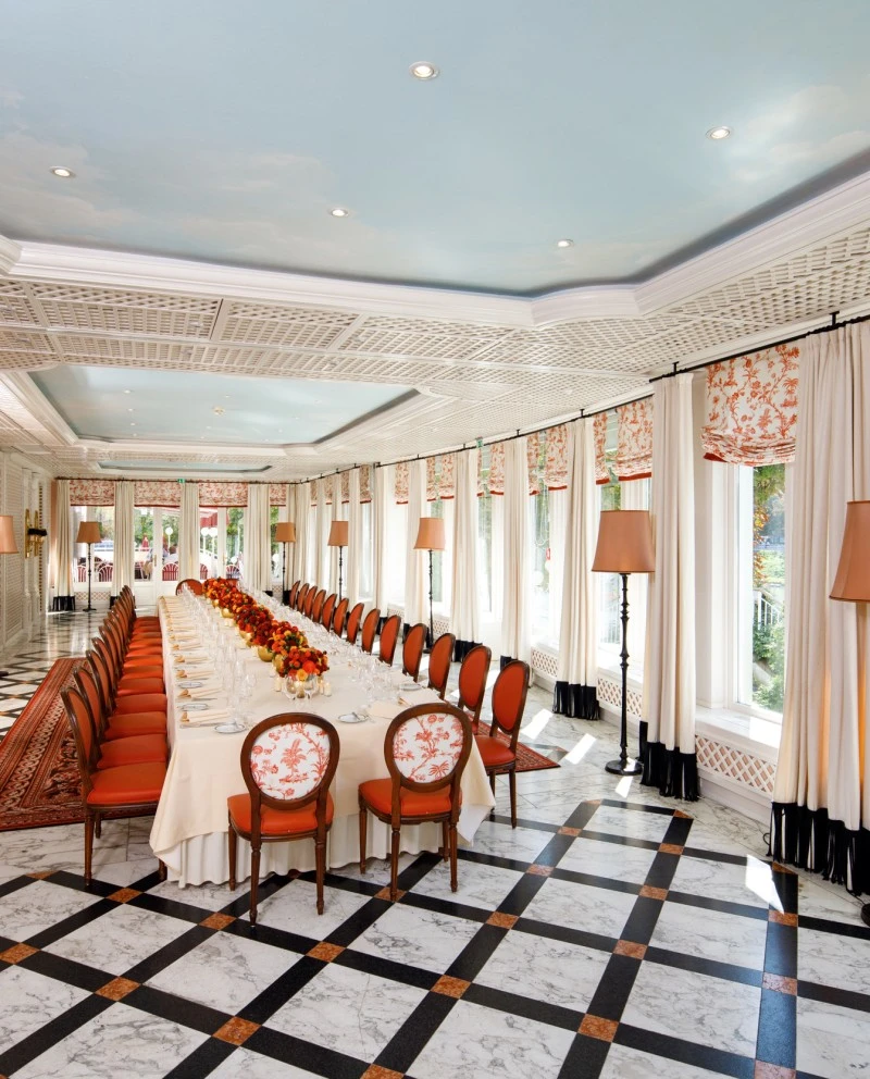 This image features a long banquet table set for a formal meal, with elegant orange chairs, floral centerpieces, and large windows that fill the bright, ornate room with natural light.