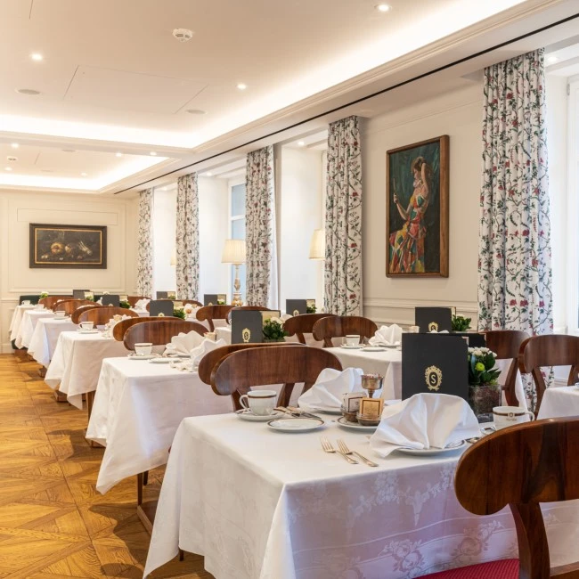 This image shows a refined dining room with neatly set tables, white linens, floral curtains, and elegant menus, ready for breakfast or a formal meal.
