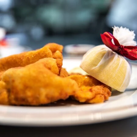 This image shows a beautifully plated Wiener Schnitzel, golden and crispy, served with a wrapped lemon wedge tied with a red ribbon for an elegant touch.