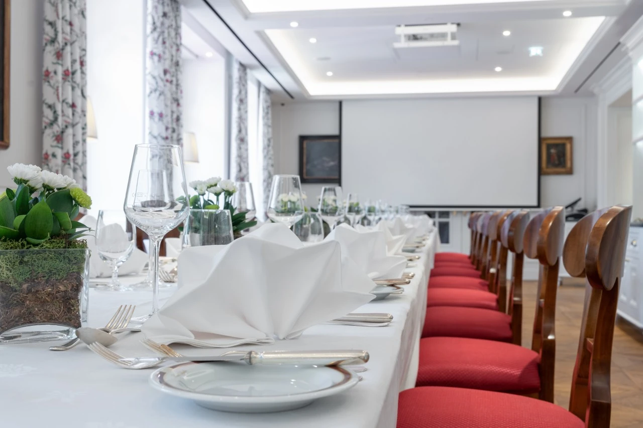 This image shows a long table elegantly set with white napkins, glassware, and floral decor, in a bright room with red chairs and a projector screen at the front.