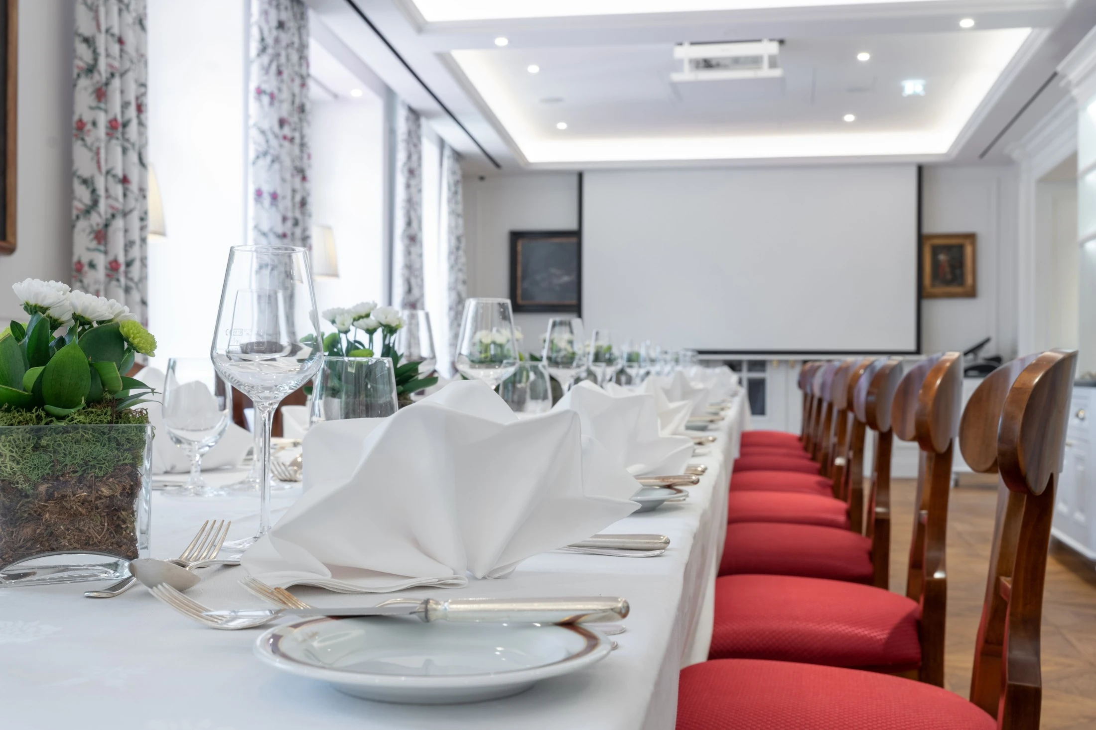 This image shows a long table elegantly set with white napkins, glassware, and floral decor, in a bright room with red chairs and a projector screen at the front.