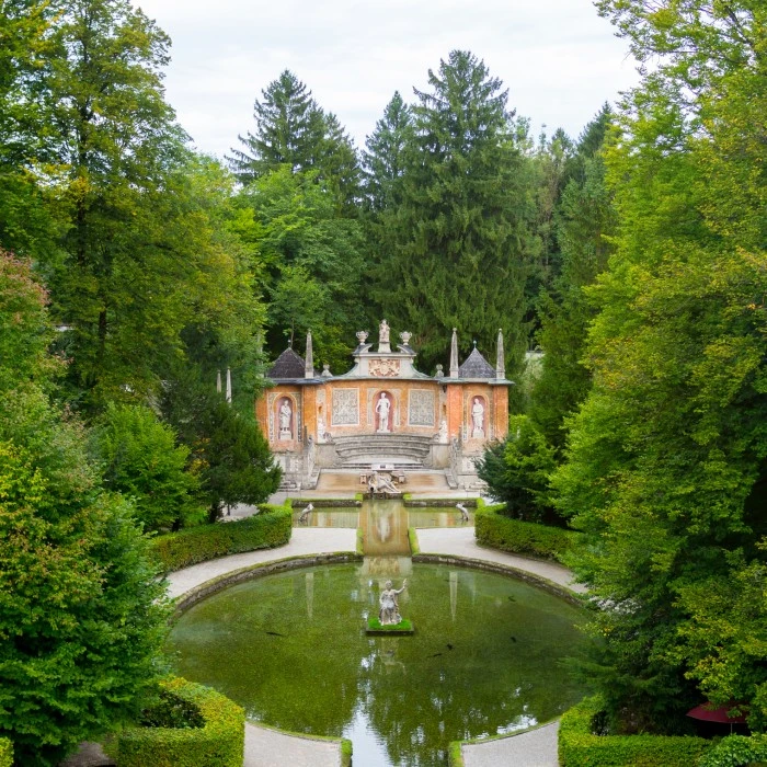 This image depicts the ornate and theatrical Roman Theatre grotto at Hellbrunn Palace, surrounded by lush forest and reflected in a circular pool with classical statues.
