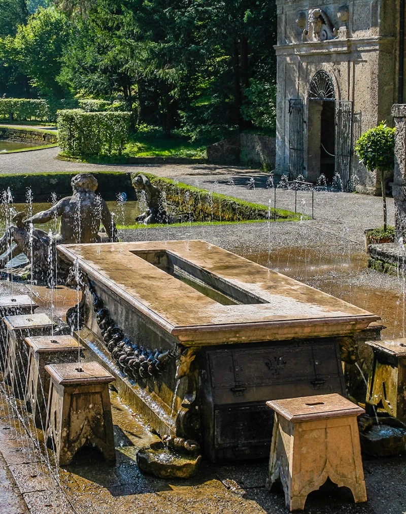 This image shows a playful trick fountain table surrounded by stools at Hellbrunn Palace in Salzburg, Austria, designed to surprise guests with hidden water jets in a beautifully manicured garden setting.