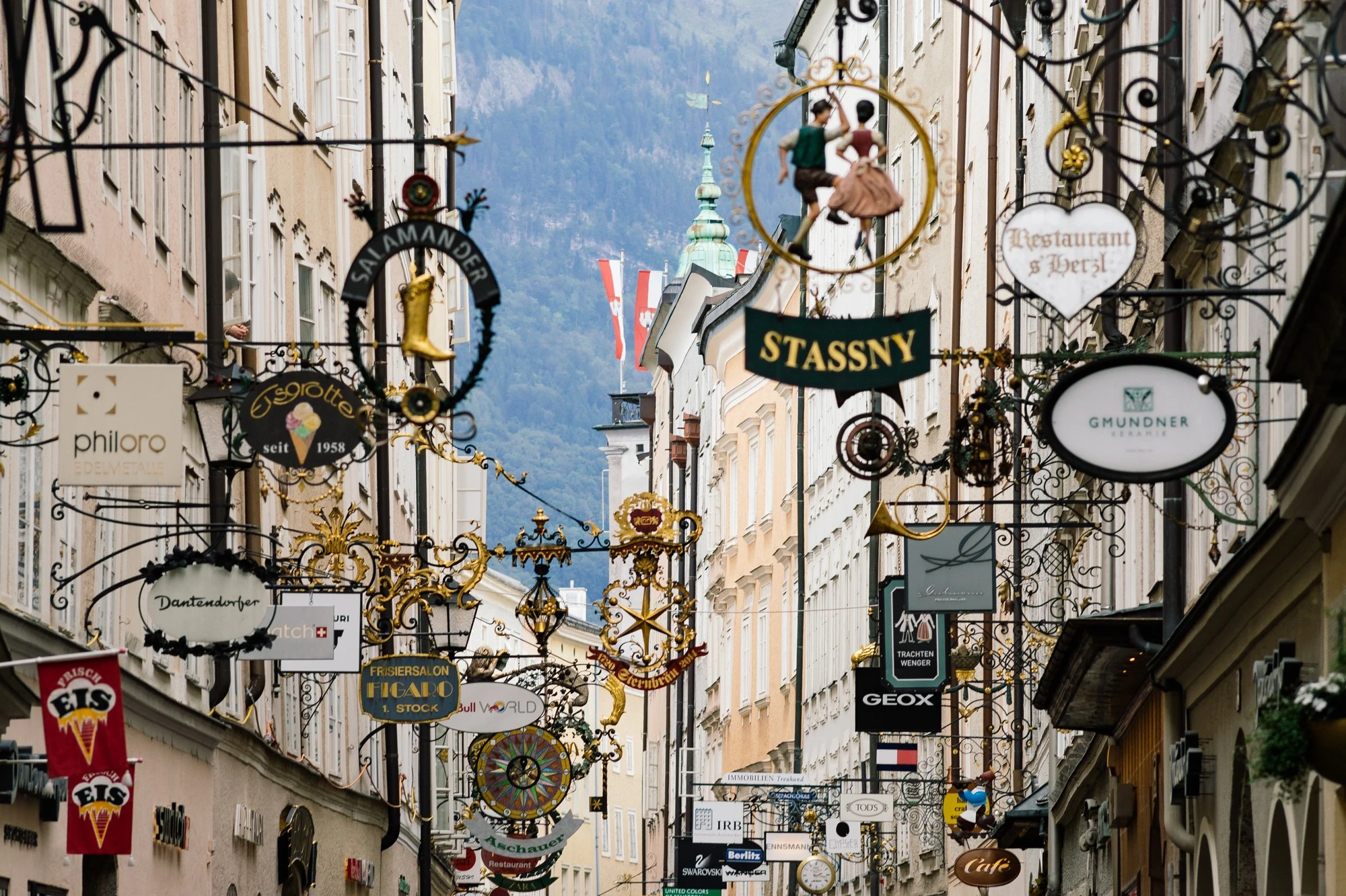 This image captures Salzburg's famous Getreidegasse, a charming historic street lined with ornate wrought-iron shop signs and flanked by old buildings set against a mountainous backdrop.