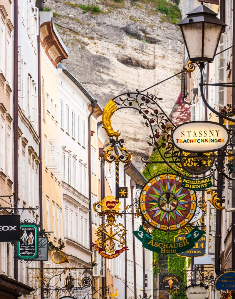 This image captures Salzburg's famous Getreidegasse, a charming historic street lined with ornate wrought-iron shop signs and flanked by old buildings set against a mountainous backdrop.