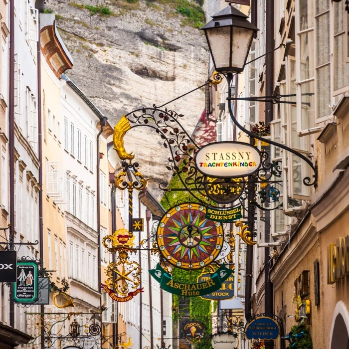 This image captures Salzburg's famous Getreidegasse, a charming historic street lined with ornate wrought-iron shop signs and flanked by old buildings set against a mountainous backdrop.