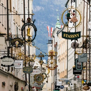 This image captures Salzburg's famous Getreidegasse, a charming historic street lined with ornate wrought-iron shop signs and flanked by old buildings set against a mountainous backdrop.