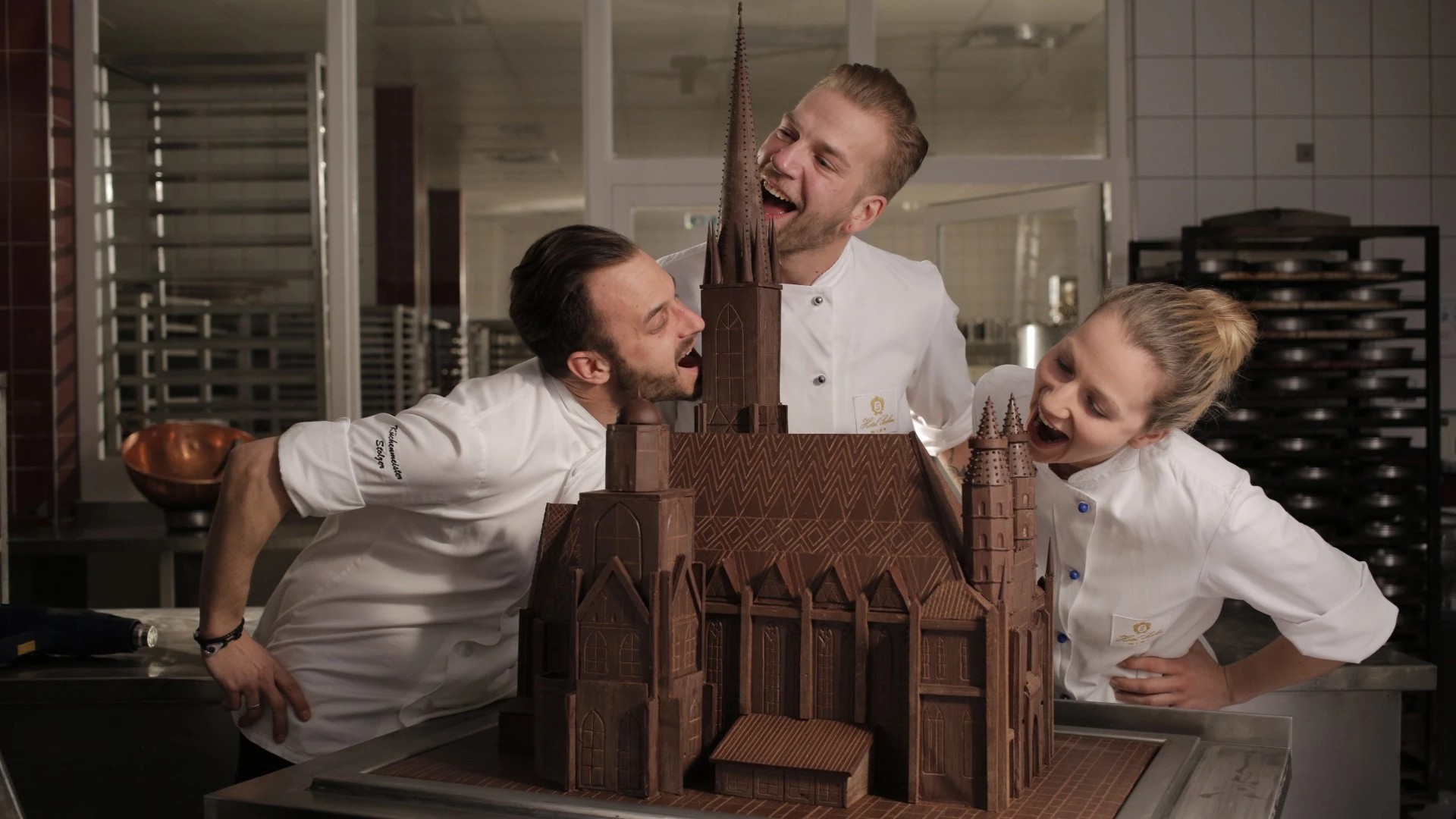 Three pastry chefs playfully pretend to bite into an intricate chocolate replica of the St.Stephens cathedral, showcasing their craftsmanship and humor in a professional kitchen.