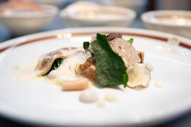 This exquisite gourmet dish is artfully plated with fresh truffle, crisp greens, delicate foam, and a medley of textures that showcase fine dining elegance at its peak.