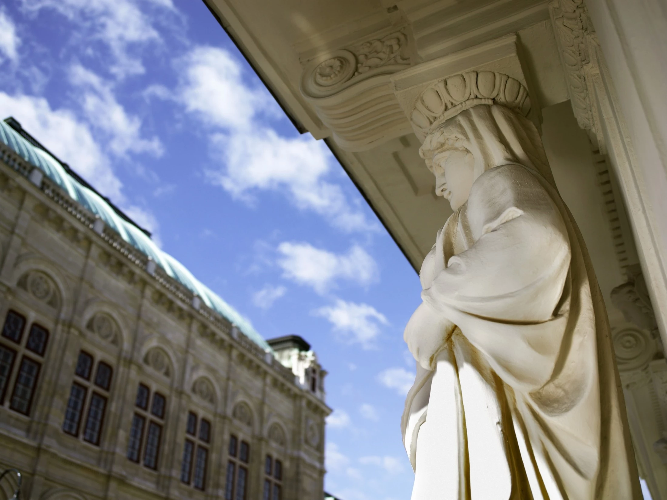 This elegant stone figure gazes thoughtfully over Vienna, adorning the exterior of Hotel Sacher with timeless grace, while the nearby Vienna State Opera rises majestically in the background under a bright blue sky.