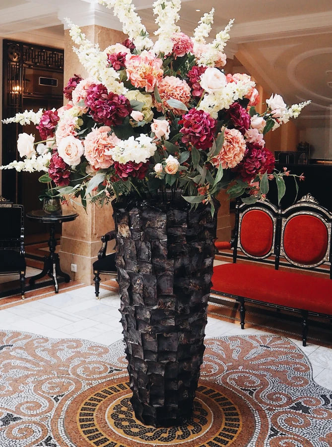 Lobby II A grand floral arrangement of blush roses, hydrangeas, and white snapdragons overflows from a striking textured vase, creating an opulent centerpiece on an intricate mosaic floor in the luxurious lobby of Hotel Sacher.