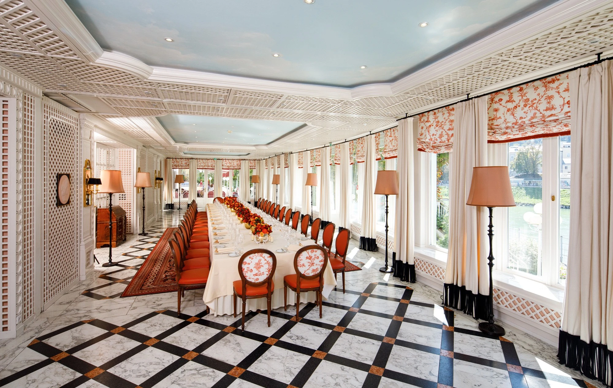 Prunkvoller Speisesaal mit langem, festlich gedecktem Tisch, flankiert von eleganten Stühlen mit orange-rotem Polster und klassischem Muster.