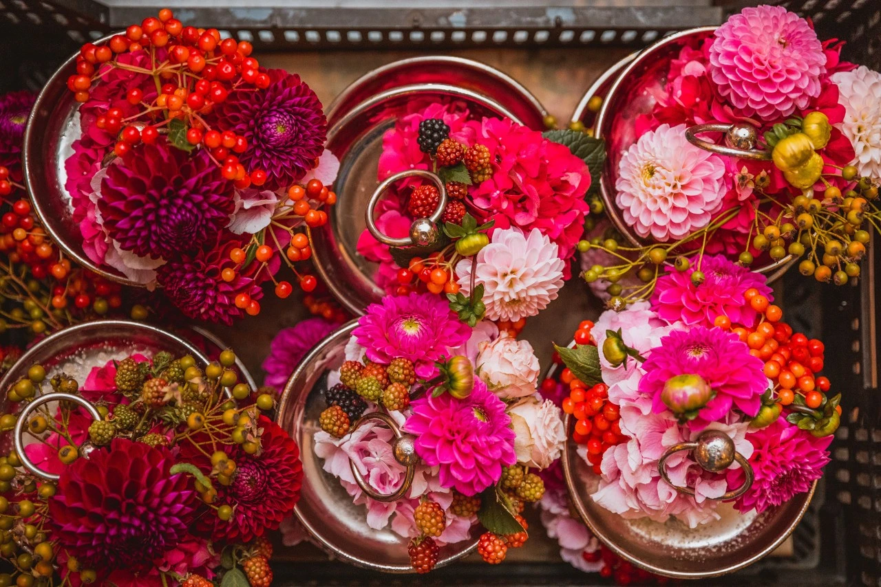 Mehrere kleine Silbergefäße, üppig gefüllt mit farbenfrohen Blüten in Pink-, Magenta- und Rosatönen sowie dekorativen Beeren in Rot, Orange und Grün. Einige Arrangements enthalten Brombeeren und glänzende silberne Kugeln, was ihnen eine festliche, luxuriöse Note verleiht.