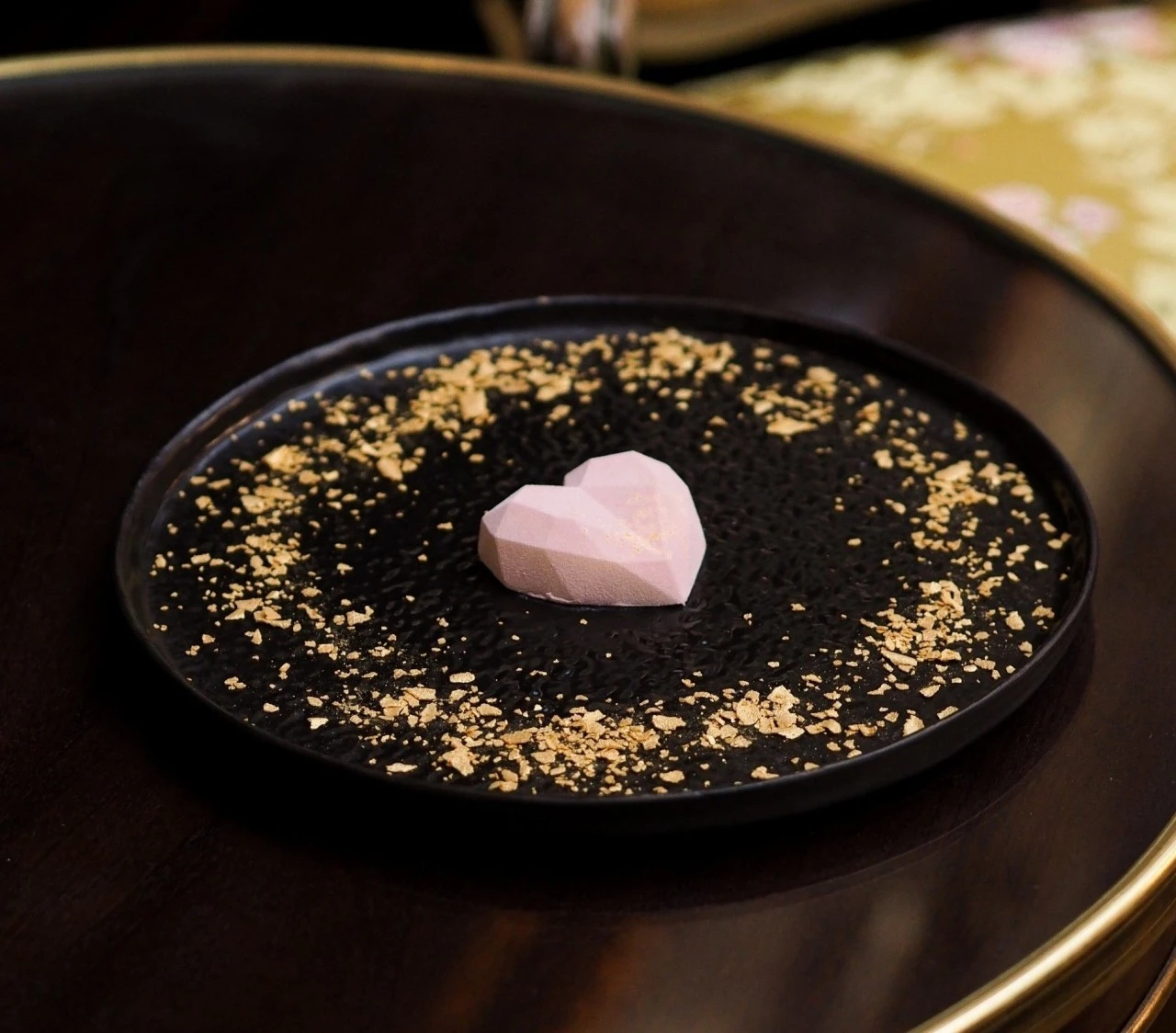 A delicate, faceted heart-shaped dessert sits at the center of a black plate dusted with shimmering gold flakes, embodying elegance, romance, and the signature luxury of Hotel Sacher.