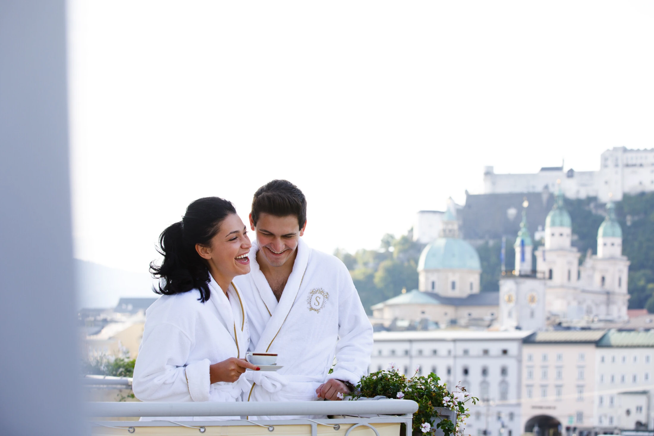 A smiling couple enjoys a peaceful morning coffee on the balcony of Hotel Sacher Salzburg, wrapped in plush robes with the historic Old Town and Hohensalzburg Fortress creating a breathtaking backdrop of timeless Austrian charm.