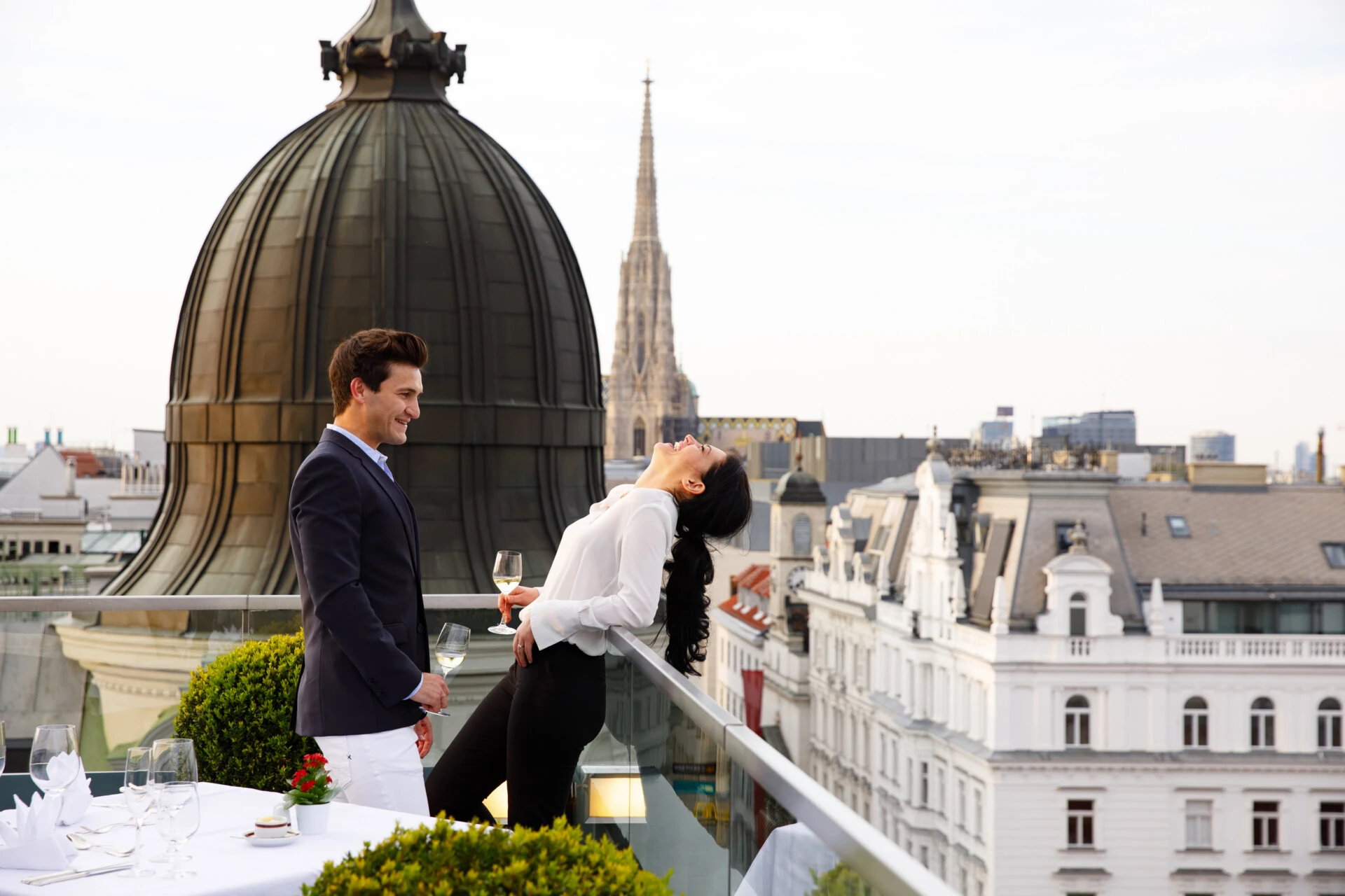 A couple shares a joyful moment with wine on the private terrace of a luxurious suite at Hotel Sacher, overlooking Vienna’s iconic skyline with St. Stephen’s Cathedral in view, capturing the essence of romance and sophistication.