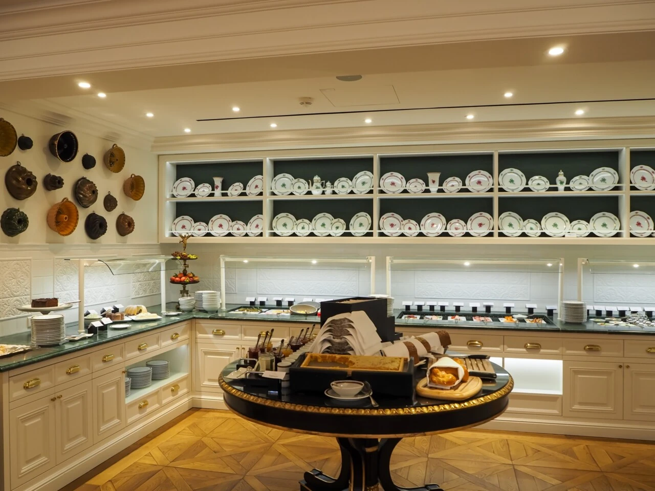 The image shows the elegant interior of Hotel Sacher Salzburg's breakfast room, featuring traditional porcelain dishes neatly arranged, polished brass cooking stations, and classic decor elements such as copper pans displayed on the wall.