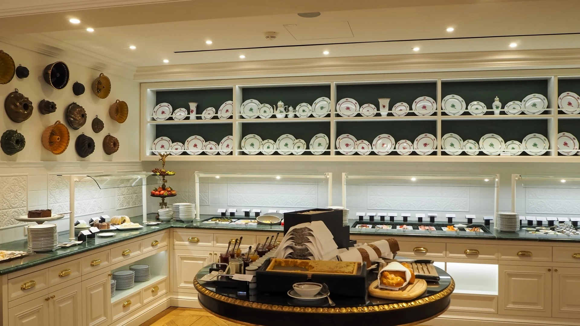 The image shows the elegant interior of Hotel Sacher Salzburg's breakfast room, featuring traditional porcelain dishes neatly arranged, polished brass cooking stations, and classic decor elements such as copper pans displayed on the wall.