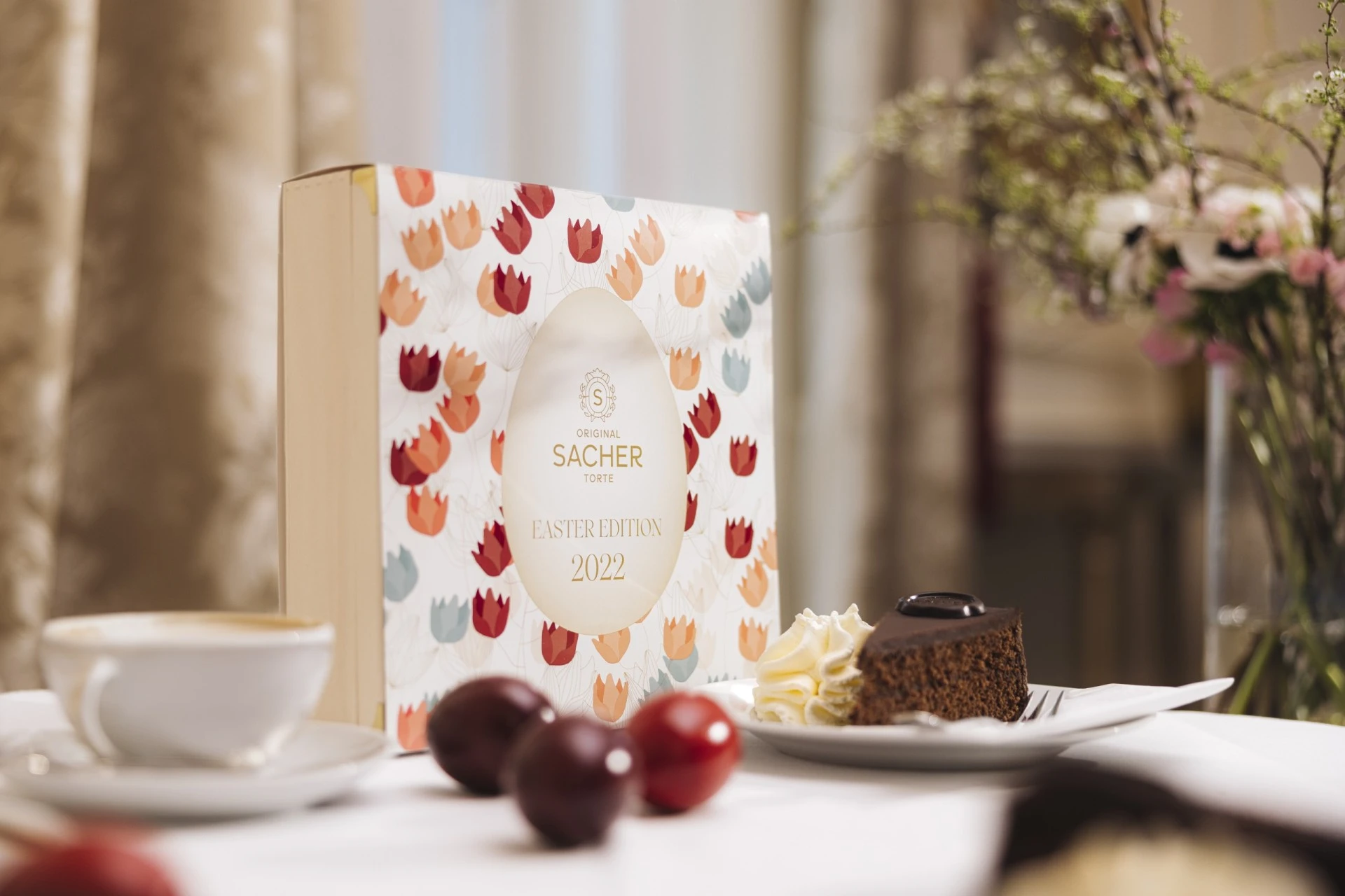 A festive table setup featuring the 2022 Easter Edition box of the Original Sacher-Torte with a delicate tulip design, accompanied by a slice of Sacher-Torte, colorful Easter eggs, and a cup of coffee, surrounded by soft spring floral decorations.