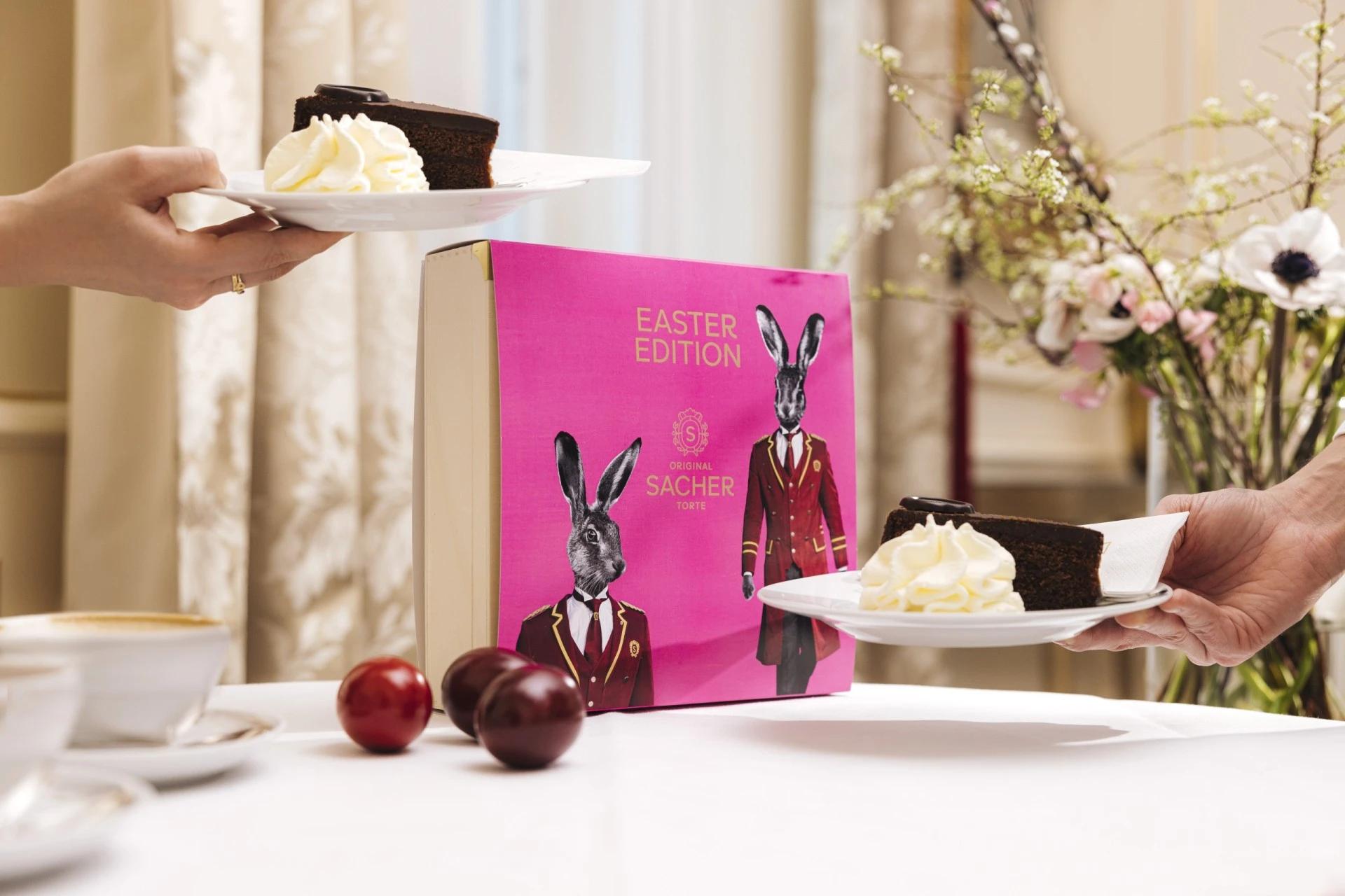 Two hands present plates with slices of the Original Sacher-Torte and whipped cream in front of the vibrant 2022 Easter Edition box, surrounded by colorful Easter eggs and spring flowers on a white tablecloth.