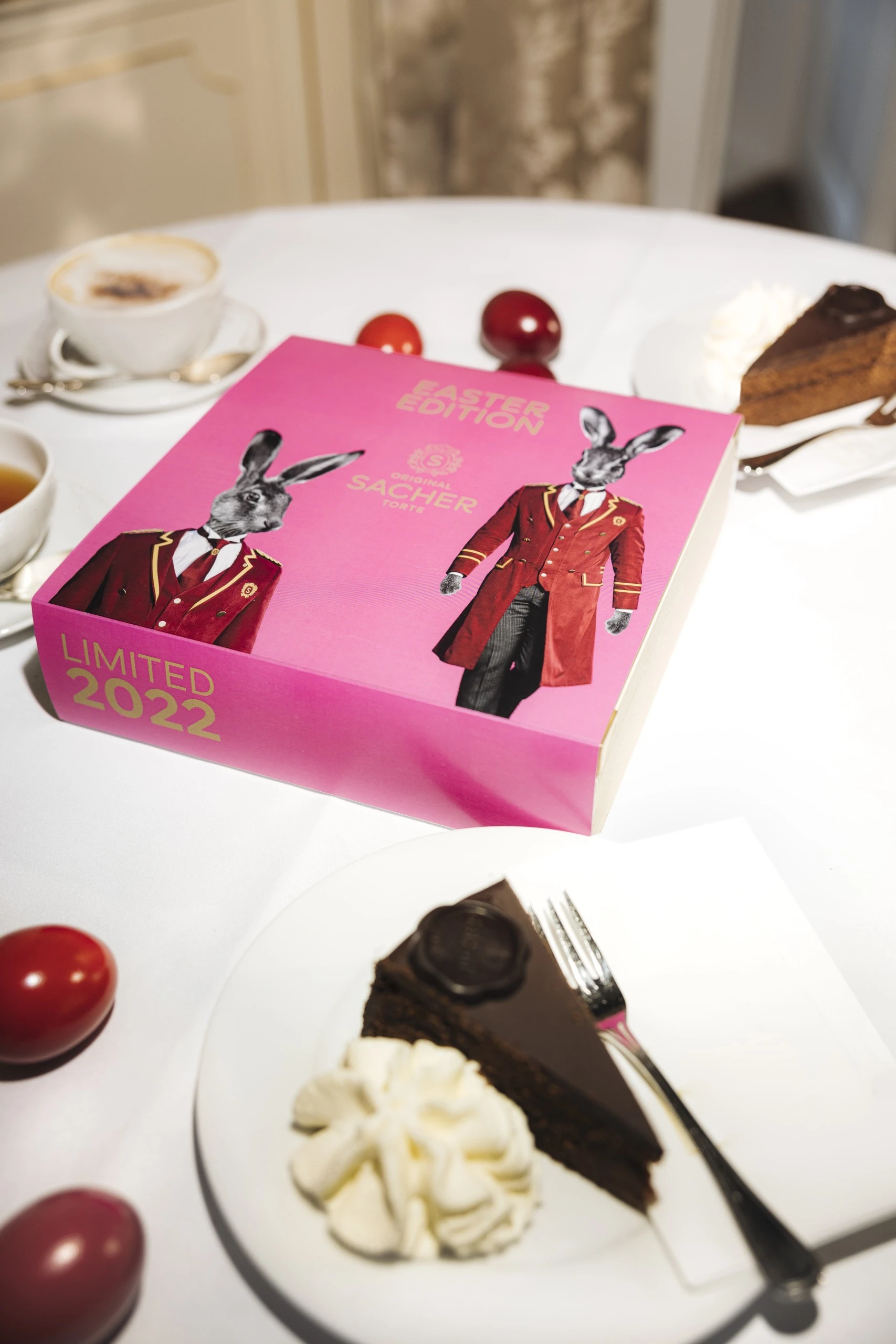 A festive table setting featuring the Limited 2022 Easter Edition box of the Original Sacher-Torte, surrounded by colorful Easter eggs, slices of Sacher-Torte with whipped cream, and cups of coffee and tea.