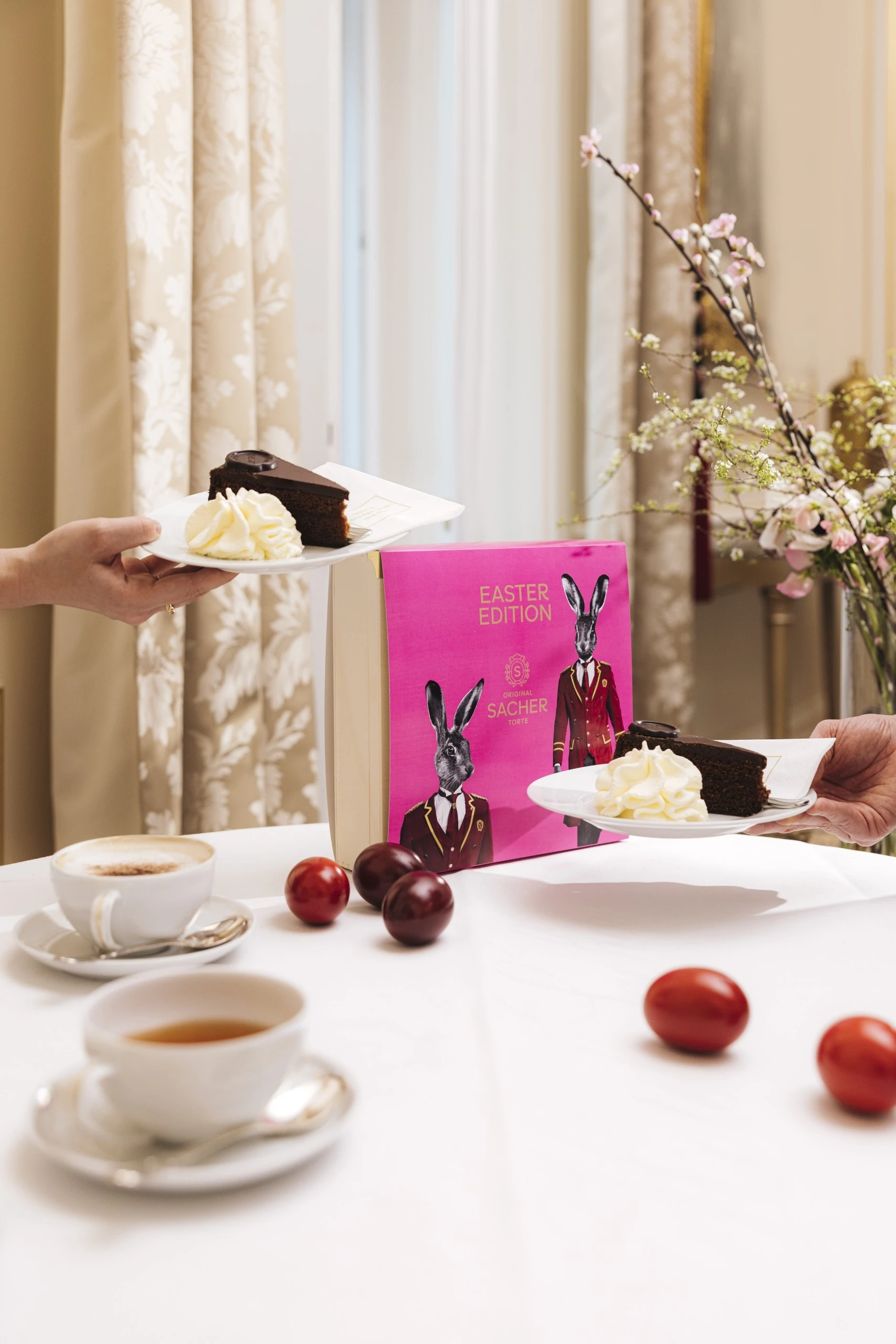 Two hands present plates with slices of Original Sacher-Torte and whipped cream in front of a vibrant pink Easter Edition box, surrounded by colorful Easter eggs and cups of coffee on a white tablecloth.