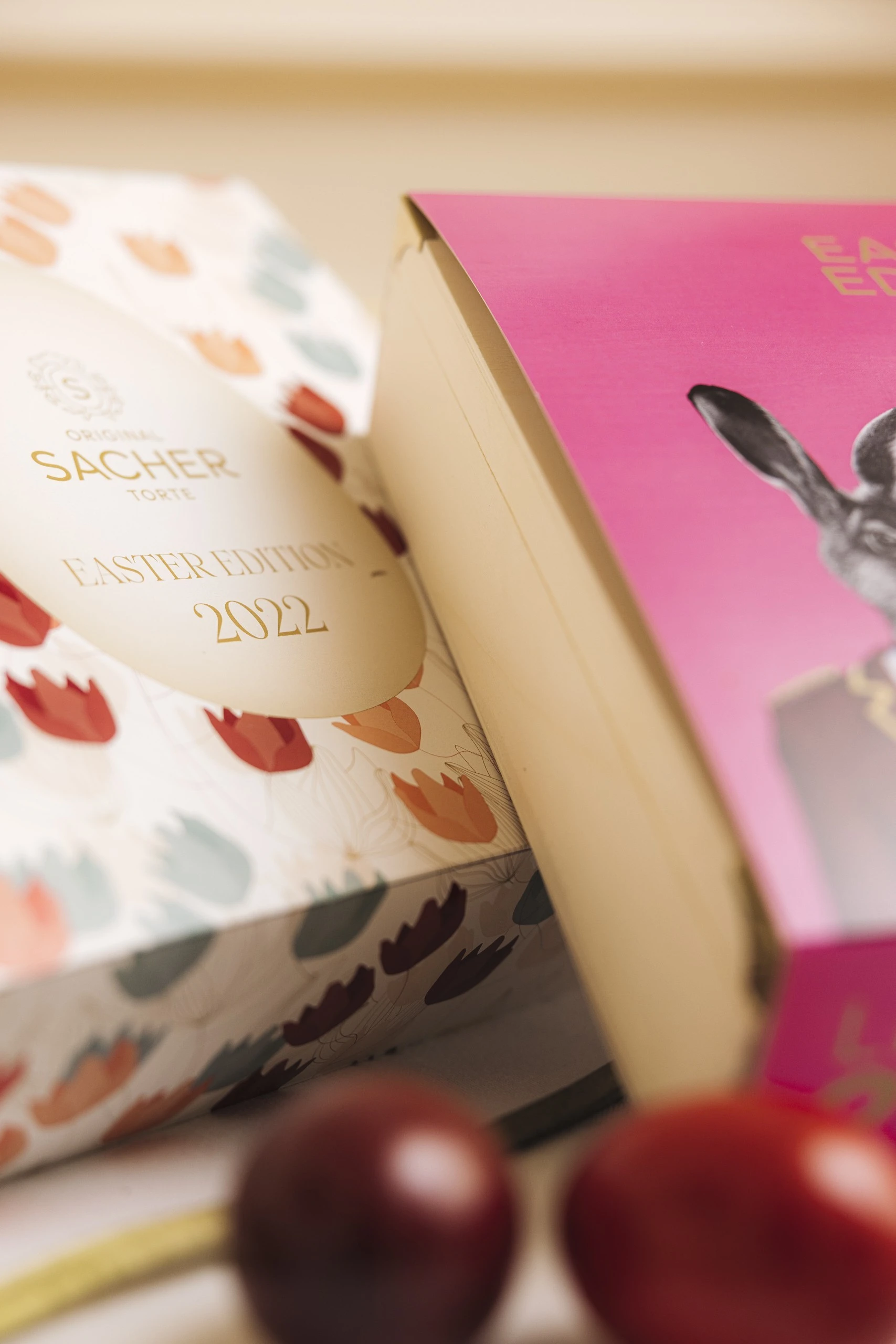 A festive arrangement featuring the Limited 2022 Easter Edition box of the Original Sacher-Torte, decorated with a rabbit illustration in a bellboy uniform, alongside a pastel floral box and colorful Easter eggs in the foreground.