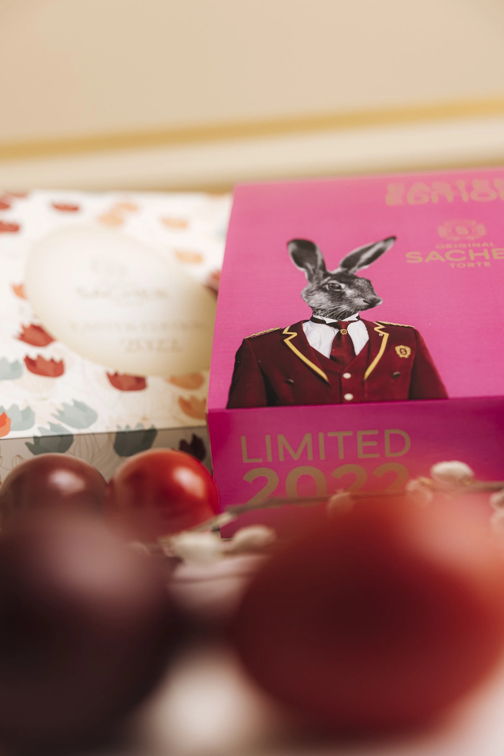 A festive arrangement featuring the Limited 2022 Easter Edition box of the Original Sacher-Torte, decorated with a rabbit illustration in a bellboy uniform, alongside a pastel floral box and colorful Easter eggs in the foreground.