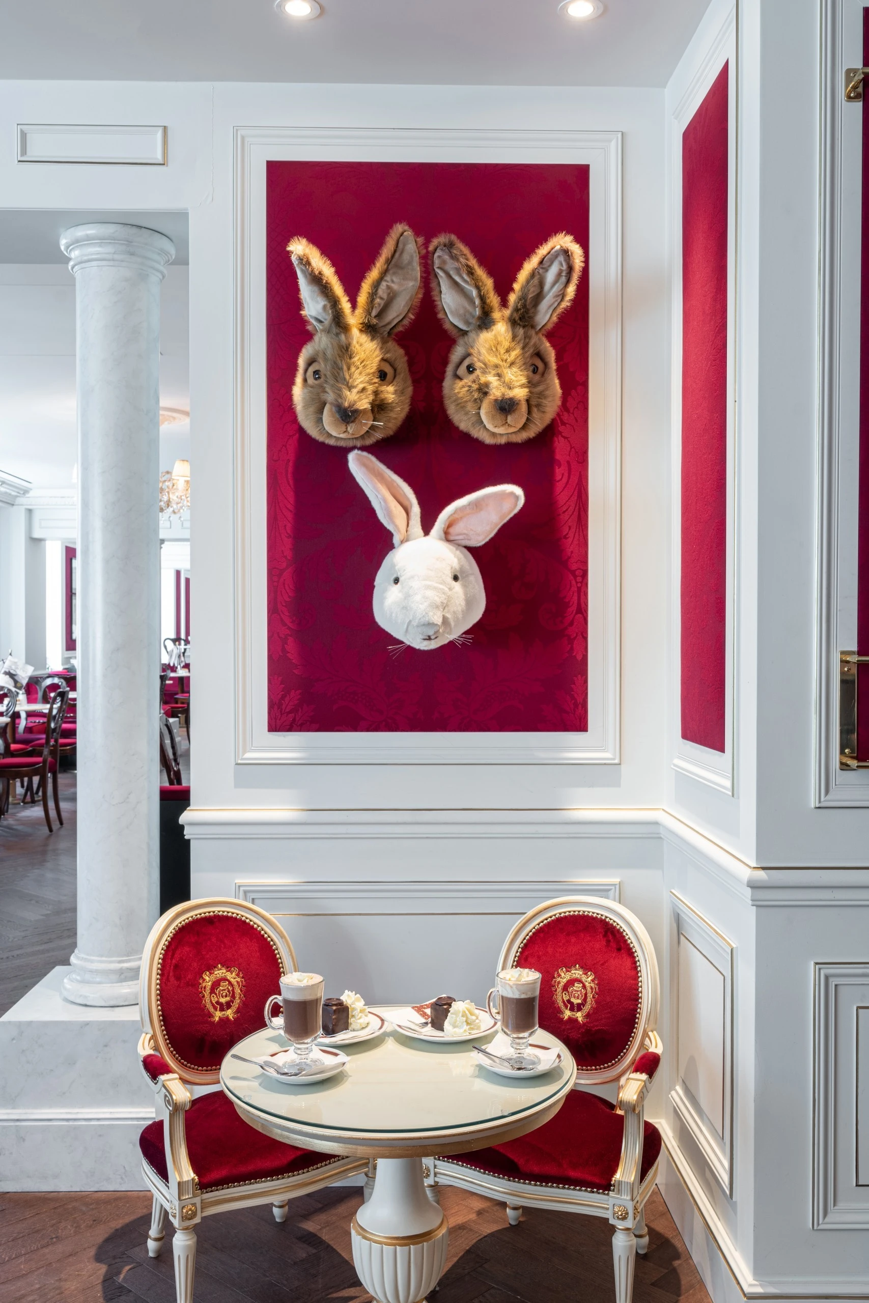A cozy table for two at Hotel Sacher, elegantly set with hot chocolate, whipped cream, and slices of Sacher-Torte, beneath a playful wall display of three rabbit heads on deep red velvet.